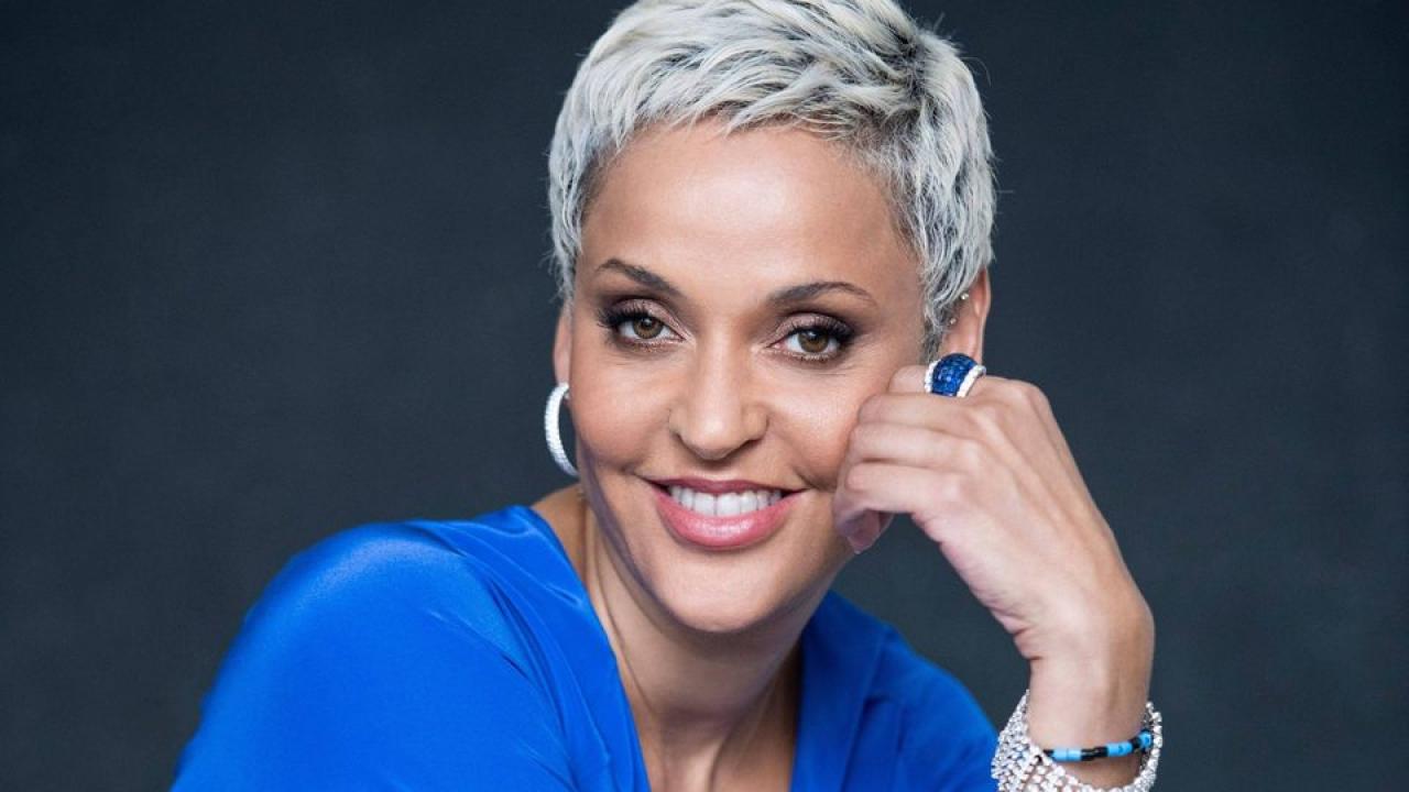Woman with silver and grey hair wearing a blue shirt smiling with their hand rested under her chin in front of a dark background