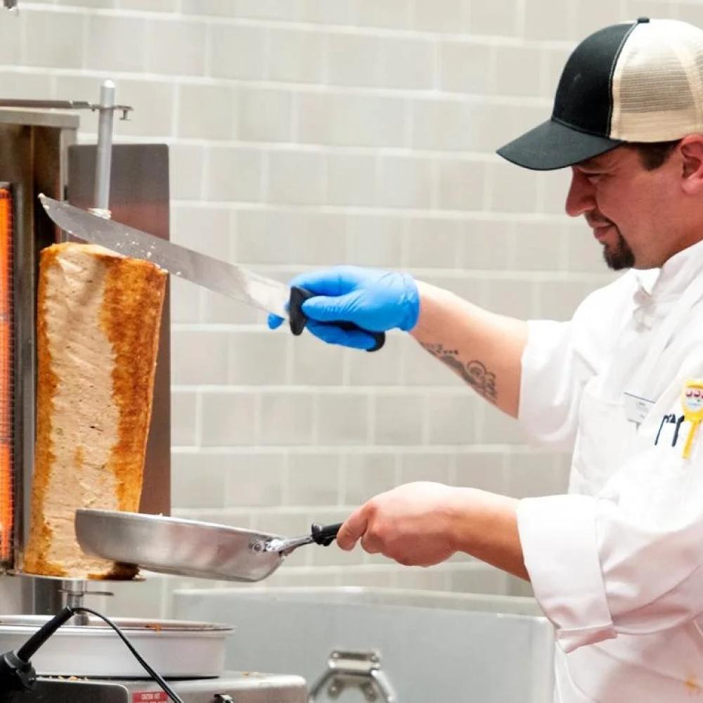 A chef in a kitchen holds a stainless steel pan and knife.