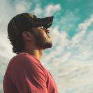 Person with eyes closed, wearing a baseball cap standing and facing toward the sun. 